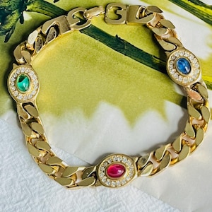 Puede incluir: Un brazalete de cadena dorada con tres piedras preciosas de forma ovalada. Las piedras preciosas son roja, azul y verde. El brazalete está sobre una superficie blanca.