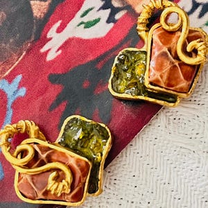 May include: A pair of gold-toned earrings with a unique design. Each earring features a textured, reddish-brown square and a green square, both framed in gold. The earrings have a decorative gold loop and swirl design.