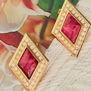 May include: A pair of gold-toned earrings in a diamond shape, each featuring a central deep magenta faceted stone surrounded by a border of small, clear, round-cut white stones set in a metallic frame with a polished finish.