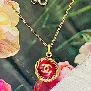 Puede incluir: Un collar dorado con un colgante rojo con un logotipo dorado. El colgante redondo tiene un borde dorado trenzado. La cadena es fina y tiene un cierre. El collar se muestra sobre un fondo floral.