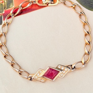 Puede incluir: Una pulsera de eslabones dorados con un adorno geométrico central engastado con una piedra roja rectangular y pequeñas piedras transparentes. La pulsera incluye un cierre metálico seguro con un elemento de diseño distintivo.