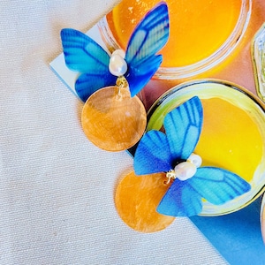 May include: A pair of earrings featuring blue butterfly charms with white pearl accents and large, round, copper-colored discs.