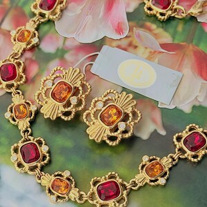 Puede incluir: Un conjunto de collar y pendientes de clip en metal dorado con cristales rectangulares facetados en rojo y naranja. Cada eslabón y componente de los pendientes está enmarcado por metal ornamentado con pequeñas piedras claras.