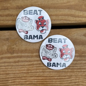 May include: Two white round buttons with the text "BEAT BAMA". Each button features a cartoon elephant and tiger, both wearing hats. The elephant has "BAMA" on its hat and "CRIMSON TIDE" on a banner. The tiger has "A" on its hat.