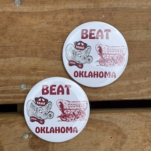 May include: Two white round buttons with the text "BEAT OKLAHOMA" in burgundy. One button features an elephant wearing a hat that says "BAMA" and the other has a covered wagon that says "SOONERS".