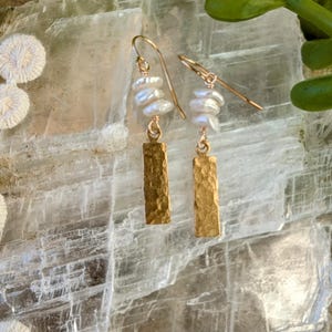 Puede incluir: Pendientes dorados con un dije rectangular martillado y un grupo de perlas blancas. Los pendientes tienen un diseño de gancho simple y se exhiben sobre una superficie texturizada y clara.