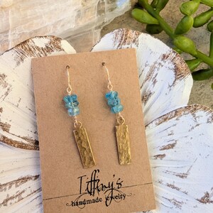 Puede incluir: Pendientes hechos a mano con rectángulos de metal martillado dorado y cuentas azul claro. Los pendientes se exhiben en una tarjeta marrón con el texto "Tiffany's handmade jewelry". El fondo incluye una flor blanca y una planta verde.