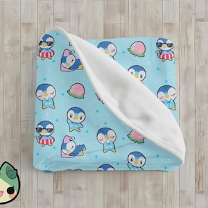 May include: A light blue blanket featuring a repeating pattern of cartoon penguins in various poses, some wearing sunglasses or hats. The blanket has a white, soft interior and is folded on a wooden surface.