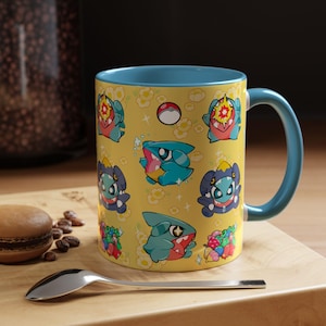 Puede incluir: Una taza de café de cerámica con un fondo amarillo con personajes de dibujos animados y motivos florales. La taza tiene un interior y un asa azules. Una cuchara y un pastel están en primer plano.