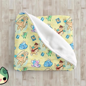 Puede incluir: Una manta amarilla con un patrón repetido de dragones de dibujos animados, criaturas azules y rosas y pequeños cuadrados azules. La manta está doblada, revelando un interior blanco. La manta está sobre una superficie de madera marrón claro.