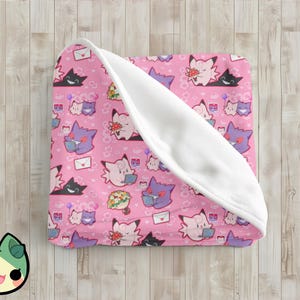 May include: Pink blanket with a repeating pattern of cartoon characters, flowers, and envelopes. The blanket is folded, revealing a white, soft interior. The blanket is on a light wood surface.