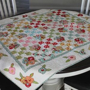May include: A white quilted table topper with a floral pattern. The design features a patchwork of squares in various colors, including red, pink, green, and blue, with floral appliqués. The border has floral designs.