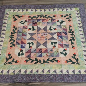 May include: A patchwork quilt with a floral design. The quilt is made of various shades of purple, pink, and green fabric. The center of the quilt features a starburst pattern. The quilt is surrounded by a border of green leaves and pink flowers.