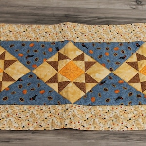 May include: A rectangular quilted table runner with a central design of three diamond-shaped star patterns. The stars are in shades of brown and orange, set against a blue background with small black bird and orange pumpkin motifs. The border is a cream color with a repeating pattern.