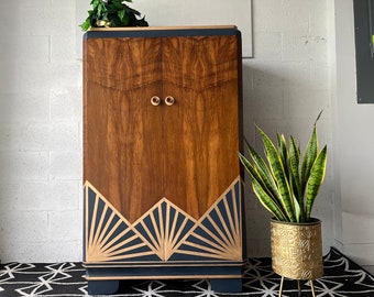 May include: A vintage wooden cabinet with two doors, painted in a dark blue color with gold accents. The cabinet has a geometric design on the bottom and brass handles. A potted plant is in the foreground.