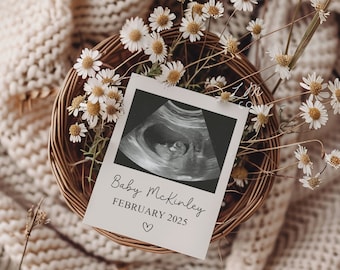 May include: A white card with the text "Baby McKinley FEBRUARY 2025" and a heart symbol. The card is placed on top of a wicker basket filled with white daisies. The basket is on a white knitted blanket.