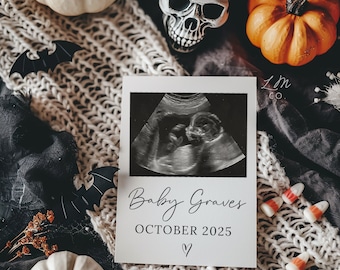 May include: A white card with a black and white ultrasound image of a baby. The card has the text "Baby Graves" and "October 2025" written in black script font. The card is surrounded by a white knitted blanket, a white pumpkin, a skull, and a small orange pumpkin.