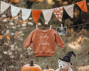 Puede incluir: Un pelele marrón con el texto "Baby Parker" y un corazón está colgado de una cuerda para tender la ropa con una hilera de banderines naranja y blancos. Dos calabazas están decoradas con las palabras "March" y "2025". Un oso de peluche con un sombrero de bruja está sentado frente a las calabazas.