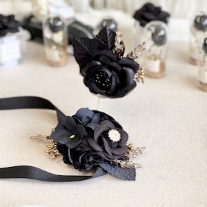 May include: Two black flower arrangements with gold accents. One arrangement is on a black ribbon and the other is on a clear glass stand.