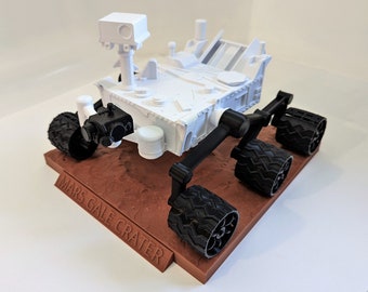 May include: A white 3D printed model of the Mars rover Curiosity, with black wheels and a red base that reads "Mars Gale Crater".