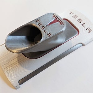 Op de afbeelding: Een 3D-geprinte Tesla Model 3 telefoonhouder in wit, grijs en rood. De houder heeft het Tesla-logo en het woord "TESLA". De telefoonhouder is ontworpen om in de middenconsole van de Model 3 te passen.