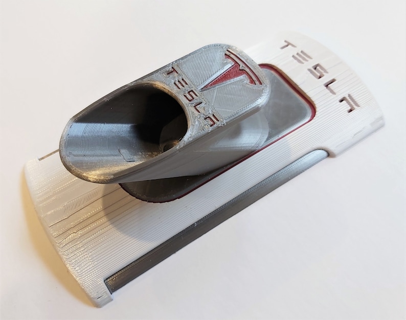 Op de afbeelding: Een 3D-geprinte Tesla Model 3 telefoonhouder in wit, grijs en rood. De houder heeft het Tesla-logo en het woord "TESLA". De telefoonhouder is ontworpen om in de middenconsole van de Model 3 te passen.