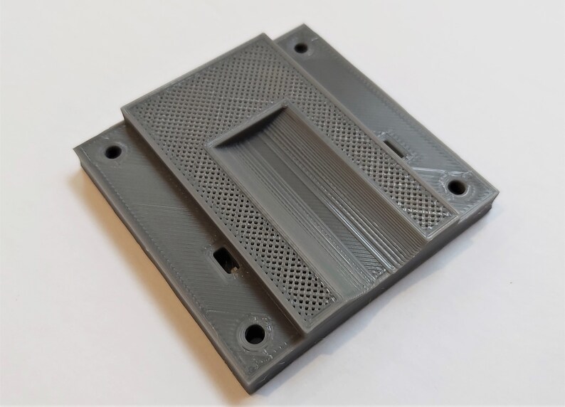 Op de afbeelding: Een grijs, 3D-geprint object met een gelaagd ontwerp. De bovenste laag heeft een geperforeerd patroon, terwijl de onderste lagen effen oppervlakken en verzonken gebieden hebben. Er zijn verschillende gaten voor montage of assemblage.