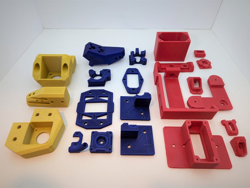 Op de afbeelding: Een verzameling 3D-geprinte onderdelen in geel, blauw en rood. De collectie bevat verschillende vormen en maten, zoals rechthoekige dozen, beugels en hoekstukken. Deze componenten lijken ontworpen te zijn voor een mechanische of technische toepassing.