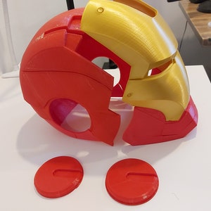 Op de afbeelding: Een gedemonteerde rood-gouden Iron Man-helm, waarschijnlijk 3D-geprint. De helm bestaat uit verschillende delen, met het voorpaneel en de oordopjes apart. De gouden secties bevinden zich op het bovenste gezicht en de rode secties vormen het onderste gezicht en de achterkant.
