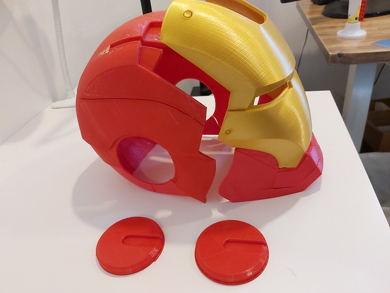 Op de afbeelding: Een gedemonteerde rood-gouden Iron Man-helm, waarschijnlijk 3D-geprint. De helm bestaat uit verschillende delen, met het voorpaneel en de oordopjes apart. De gouden secties bevinden zich op het bovenste gezicht en de rode secties vormen het onderste gezicht en de achterkant.