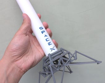 May include: A white SpaceX rocket model with blue lettering, held in a hand. The rocket has a grey landing structure at the base. The model is a miniature replica of a spacecraft.