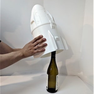Könnte beinhalten: Ein weißer, 3D-gedruckter Flaschenverschluss in Form einer Rakete, der auf eine Weinflasche gelegt wird.