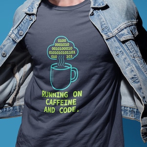 Può includere: T-shirt grigio scuro con grafica verde neon di una tazza di caffè e una nuvola di codice binario. Il testo "RUNNING ON CAFFEINE AND CODE" è sotto. La persona indossa una giacca di jeans.
