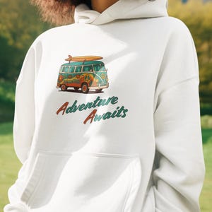 Könnte beinhalten: Weißer Kapuzenpullover mit einer Vordertasche. Er zeigt eine farbenfrohe Illustration eines Oldtimer-Vans mit einem Surfbrett. Der Text "Adventure Awaits" ist darunter in Rot und Grün gedruckt.