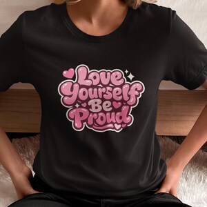Può includere: T-shirt nero con la scritta "Love Yourself Be Proud" in un design a lettere a bolle. Il testo è rosa e bianco con dettagli a forma di cuore e stella. La maglietta è a girocollo.