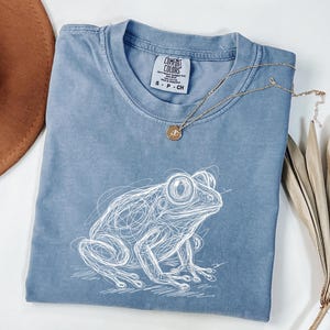 May include: A blue Comfort Colors t-shirt with a white sketch of a frog. The shirt has a crew neck and short sleeves. A gold necklace with a small pendant is draped across the shirt. A brown hat and dried plant are in the background.