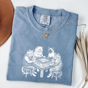 May include: A blue Comfort Colors t-shirt featuring a white illustration of four cats playing a game at a table. The cats are anthropomorphic, dressed in clothing, and seated around a table with game pieces. A gold necklace with a pendant is draped on the shirt.