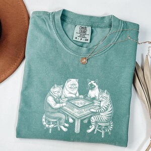 May include: A teal Comfort Colors t-shirt featuring a graphic of four cats playing a board game. The cats are drawn in a detailed, cartoon style. A silver necklace with a small pendant is draped across the shirt.