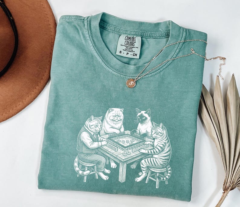 May include: A teal Comfort Colors t-shirt featuring a graphic of four cats playing a board game. The cats are drawn in a detailed, cartoon style. A silver necklace with a small pendant is draped across the shirt.
