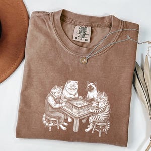 May include: A brown Comfort Colors t-shirt featuring a whimsical illustration of four cats playing a board game. The cats are depicted in a detailed, cartoon style, with one wearing a vest. A silver necklace with a small pendant is draped across the shirt.
