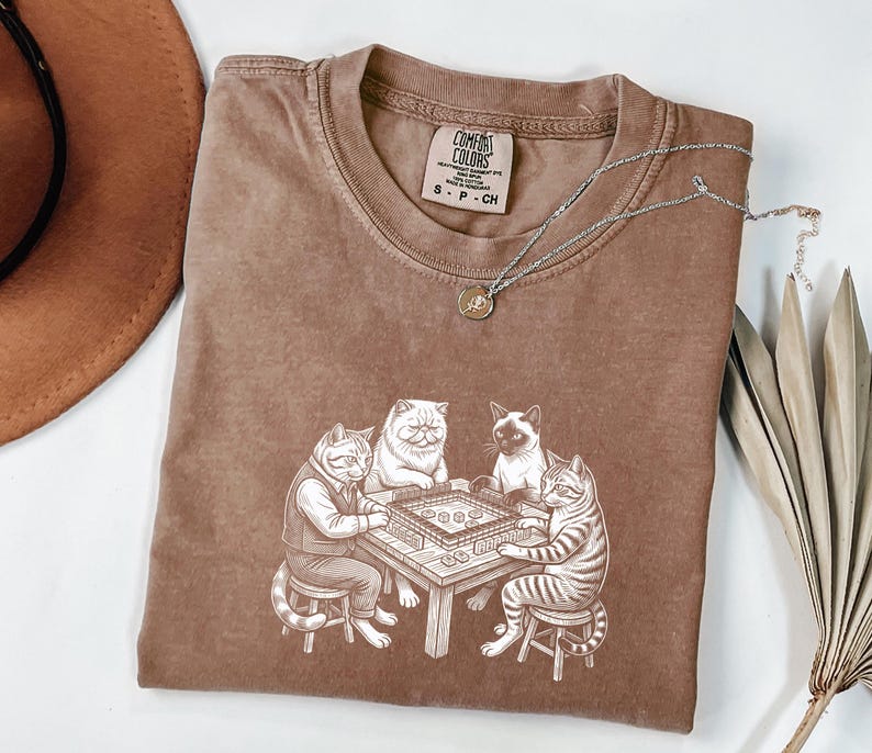May include: A brown Comfort Colors t-shirt featuring a whimsical illustration of four cats playing a board game. The cats are depicted in a detailed, cartoon style, with one wearing a vest. A silver necklace with a small pendant is draped across the shirt.