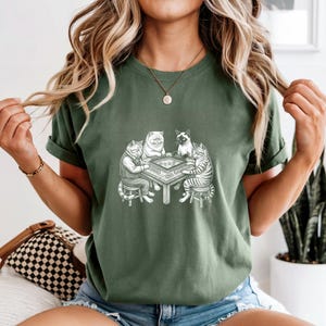 May include: Green t-shirt featuring a detailed illustration of four cats playing a board game around a table. The cats are depicted in various poses, with one wearing a jacket. The design is in black and white.