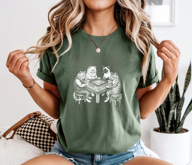 May include: Green t-shirt featuring a detailed illustration of four cats playing a board game around a table. The cats are depicted in various poses, with one wearing a jacket. The design is in black and white.