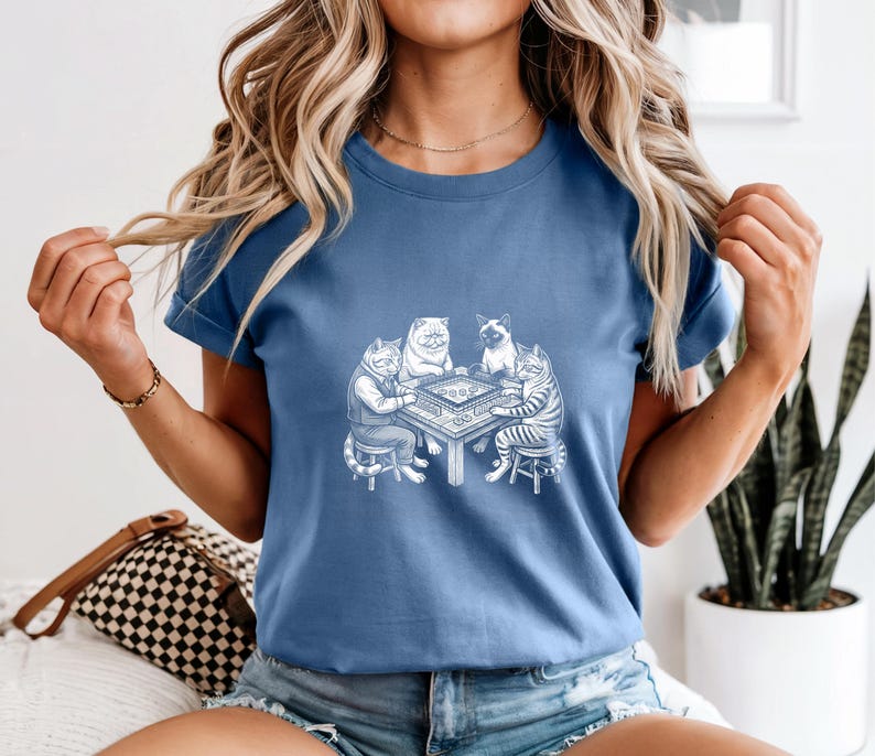 May include: A blue t-shirt featuring a white illustration of four cats playing a board game. The cats are anthropomorphic, dressed in clothing, and seated around a table. The shirt is made of a soft material.