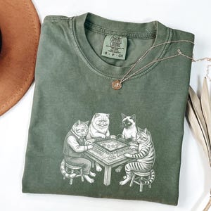 May include: Olive green Comfort Colors t-shirt featuring a graphic of four cats playing a game at a table. The cats are drawn in a detailed, cartoon style. A gold necklace with a small pendant rests on the shirt. The shirt is size S-P-CH.