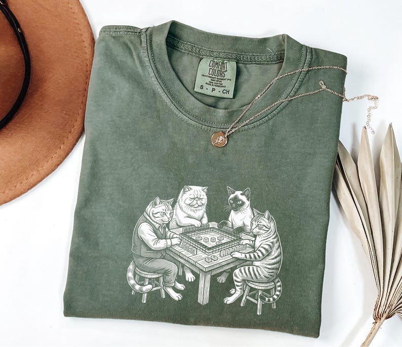 May include: Olive green Comfort Colors t-shirt featuring a graphic of four cats playing a game at a table. The cats are drawn in a detailed, cartoon style. A gold necklace with a small pendant rests on the shirt. The shirt is size S-P-CH.