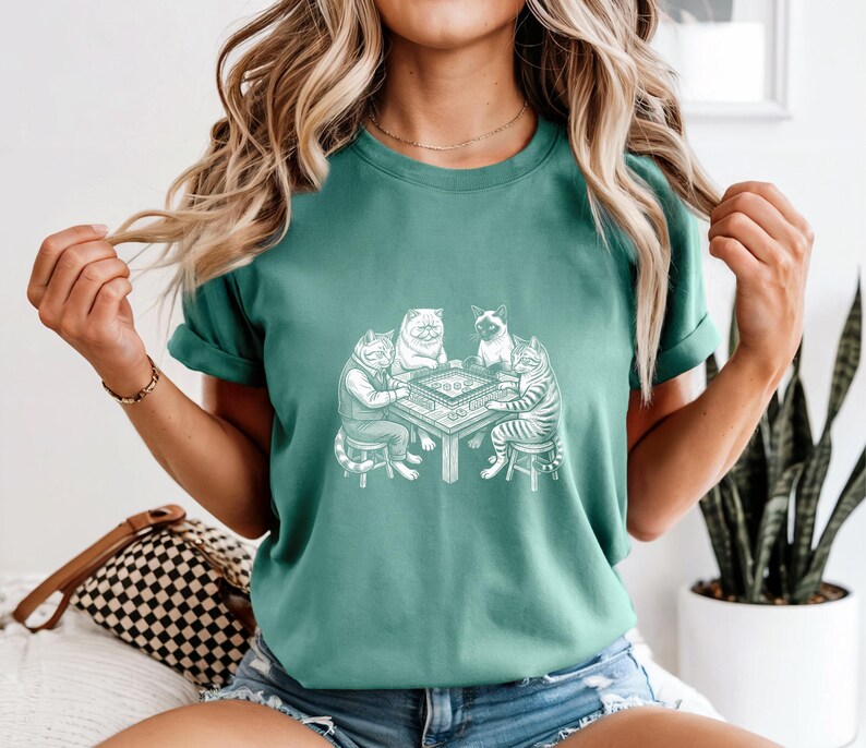 May include: Teal-colored t-shirt featuring a detailed graphic of four cats playing a board game. The cats are anthropomorphic, dressed in clothing, and seated around a table. The graphic is in a detailed, line-art style, creating a whimsical and unique design.