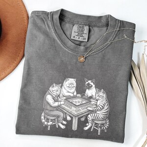 May include: A gray Comfort Colors t-shirt featuring a graphic of four cats playing mahjong. The cats are depicted in a detailed, cartoon style, with one wearing a vest. A gold necklace is draped across the shirt.