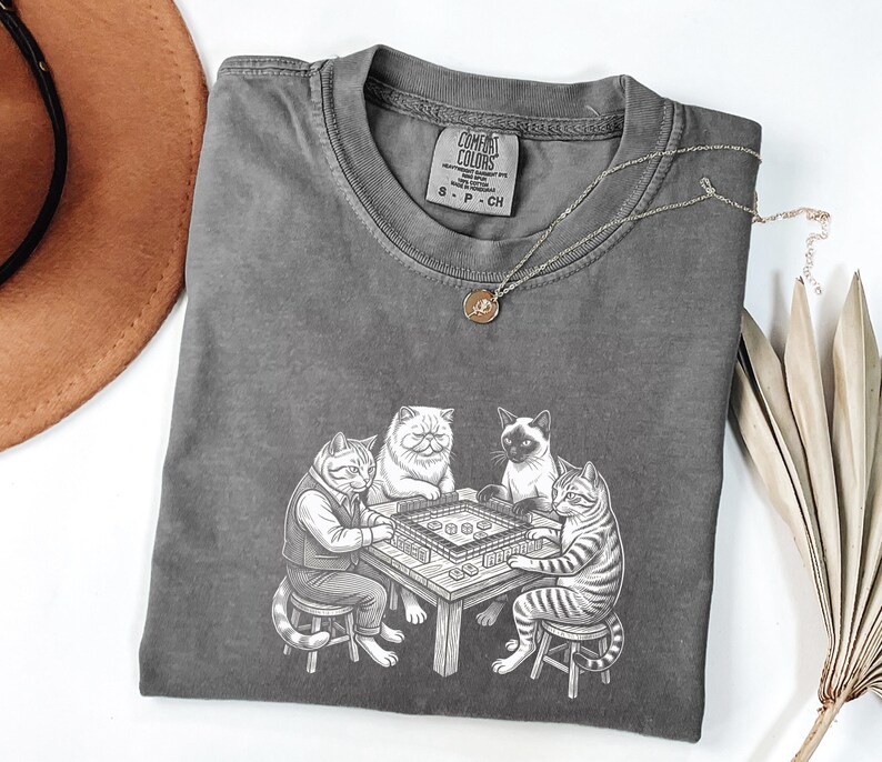 May include: A gray Comfort Colors t-shirt featuring a graphic of four cats playing mahjong. The cats are depicted in a detailed, cartoon style, with one wearing a vest. A gold necklace is draped across the shirt.