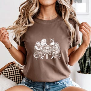 May include: A brown t-shirt featuring a detailed illustration of four cats playing a board game. The cats are depicted in a vintage style, with one wearing a vest. The shirt is a solid color and has short sleeves.